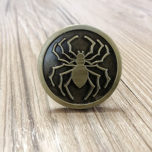 Może przedstawiać: Okrągły, złoty metalowy gałka z czarnym wzorem pająka.