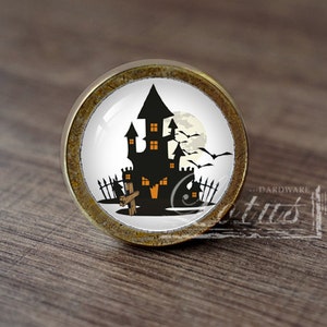 Pode incluir: Um botão de armário redondo de cor latão com um inserto de cerâmica branca que apresenta uma silhueta preta de uma casa assombrada com lua cheia e morcegos. A casa tem janelas laranja e uma cruz na frente.