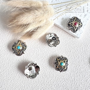 Puede incluir: Botones decorativos de metal plateado y dorado, con detalles de piedras preciosas turquesas y rojas. Los botones tienen un diseño geométrico intrincado. Algunos están sobre una superficie blanca con hierba ornamental seca.