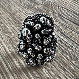 K&ouml;nnte beinhalten: Ein silberner Ring mit einem Design aus zahlreichen kleinen Totenk&ouml;pfen. Der Ring hat eine strukturierte Oberfl&auml;che und einen dunklen Hintergrund. Die Totenk&ouml;pfe sind aufwendig detailliert, was dem Ring eine gotische oder alternative &Auml;sthetik verleiht.