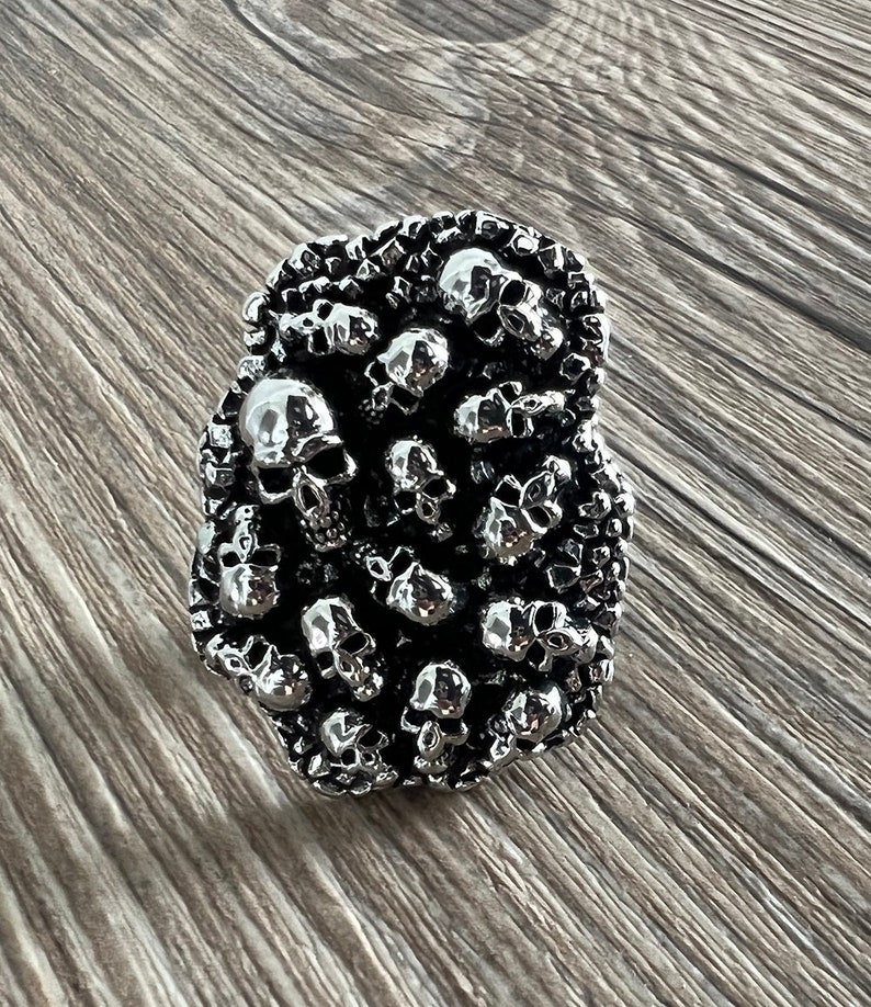 K&ouml;nnte beinhalten: Ein silberner Ring mit einem Design aus zahlreichen kleinen Totenk&ouml;pfen. Der Ring hat eine strukturierte Oberfl&auml;che und einen dunklen Hintergrund. Die Totenk&ouml;pfe sind aufwendig detailliert, was dem Ring eine gotische oder alternative &Auml;sthetik verleiht.