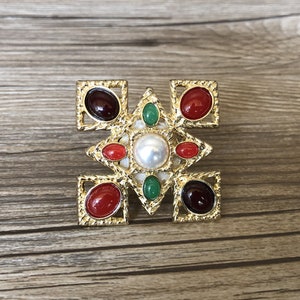May include: A gold-toned brooch with a geometric design featuring red, green, and white stones. The brooch has a central pearl surrounded by four smaller stones.