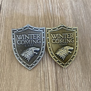 以下が含まれることがあります： 「Winter is Coming」と「Stark」の文字が書かれた、金属製の盾型のピン2つ。ピンは盾の形をしており、中央にはダイアウルフの頭が描かれている。1つのピンは銀色、もう1つは金色です。