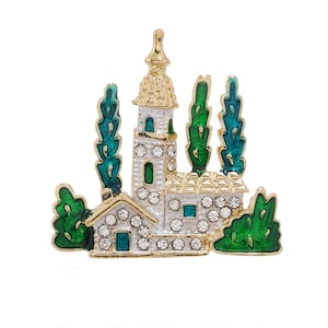 May include: A gold-tone brooch featuring a white enamel church and green enamel trees. The church has a gold-tone roof and windows with clear rhinestones. The trees are stylised and have a green enamel finish.