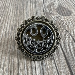 May include: A silver and bronze circular pin featuring a black and white image of a smiling face with a web-like design. The pin is surrounded by a decorative border of small flowers.