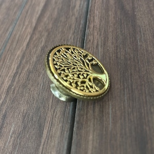 May include: A gold-toned metal cabinet knob with a tree of life design. The knob is round and has a raised design of a tree with branches and leaves.