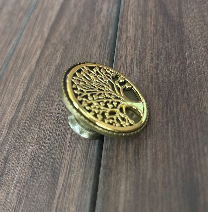 May include: A gold-toned metal cabinet knob with a tree of life design. The knob is round and has a raised design of a tree with branches and leaves.