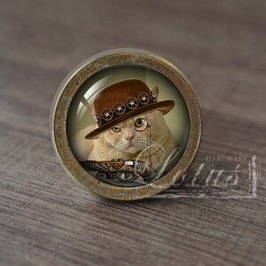 May include: A round, brass-colored cabinet knob with a glass insert featuring a cat wearing a brown cowboy hat with a gold band, a monocle, and holding a gun. The cat has a serious expression.