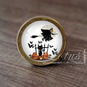 Pode incluir: Um botão de armário redondo de cor dourada com um inserto de cerâmica branca que apresenta um gato preto, uma bruxa voando em uma vassoura e morcegos contra uma lua cheia. O botão tem um aspecto desgastado e envelhecido.