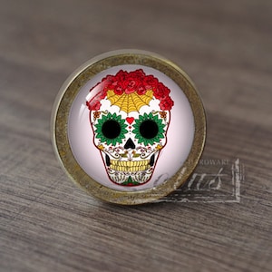 Puede incluir: Un pomo de armario redondo de color latón con un fondo blanco y un diseño de calavera de azúcar colorido. La calavera tiene ojos verdes, un corazón rojo y un patrón floral.