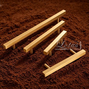 May include: Four gold-colored, rectangular cabinet pulls of varying sizes are arranged on a textured, brown surface. The pulls have a simple, modern design with a bar shape and cylindrical posts for mounting. The image has a warm, inviting tone.