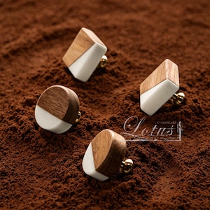 Può includere: Quattro pomelli decorativi per mobili con un design geometrico. Ogni pomello presenta una combinazione di legno, ceramica bianca e una base dorata. I pomelli sono disposti su una superficie marrone strutturata.