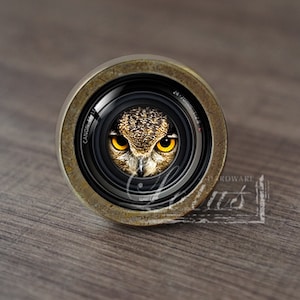 Puede incluir: Un pomo de lente de cámara redondo de color latón con la cara de un búho en el centro. El búho tiene ojos amarillos y plumas marrones. La lente está etiquetada con "CANON" y "EF 50mm 1:1.8".