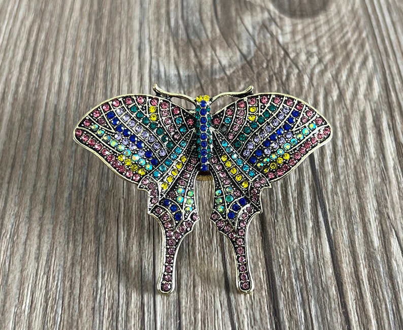 May include: A colourful butterfly brooch with a gold-tone finish. The butterfly is covered in iridescent rhinestones in shades of pink, blue, green, and yellow.