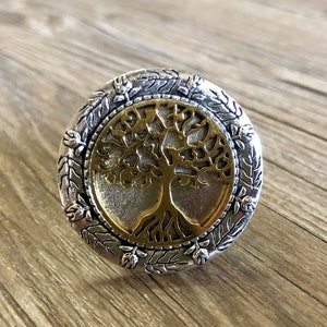 May include: A silver ring featuring a gold tree of life design. The ring is round and has a detailed border of leaves and flowers.