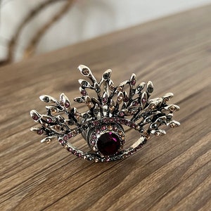 以下が含まれることがあります： 中央に紫色の宝石が埋め込まれたシルバーリング。宝石の周りを小さなラインストーンが囲んでいます。リングは葉のデザインで囲まれた目の形をしています。