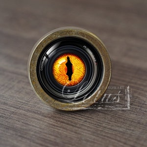 Puede incluir: Primer plano de un pomo redondo de color latón con una lente de cámara negra en el centro. La lente tiene un ojo naranja y amarillo brillante en su interior.