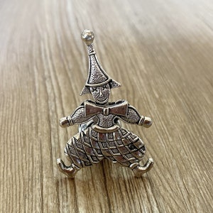 Op de afbeelding: Een zilverkleurige metalen servetring in de vorm van een nar met een puntige hoed, een strik en een geruit patroon op de broek.