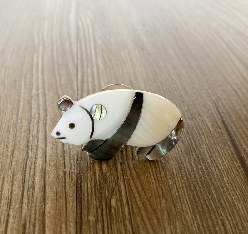 Puede incluir: Un broche de panda blanco y negro hecho de n&aacute;car con acentos iridiscentes.