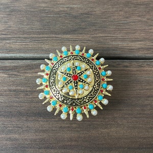 May include: A gold-toned brooch with a circular design featuring a starburst pattern with red, turquoise, and white stones. The brooch is surrounded by a ring of white pearls and gold spikes.