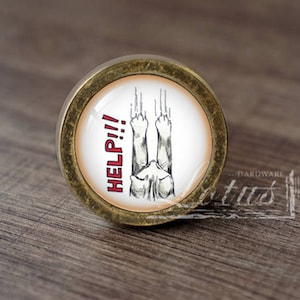 Może przedstawiać: Okrągły, złoty gałka meblowa z białym tłem i czarno-białym rysunkiem kota z uniesionymi łapami, który mówi "HELP!!!" czerwonymi literami.