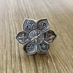 May include: Silver ring with a flower design featuring symbols of different religions, including a cross, a star, a peace sign, and a yin yang symbol.
