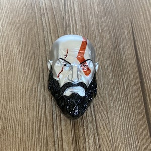 May include: A silver and black figurine of a man with a beard and red markings on his face. The figurine is a representation of the Greek god Kratos.