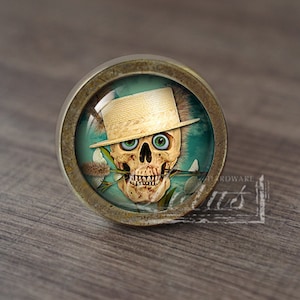 Pode incluir: Um botão de armário redondo de cor latão com um inserto de vidro que apresenta uma caveira com um chapéu de palha e segurando uma flor entre os dentes. O fundo é azul turquesa.