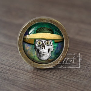 Pode incluir: Um botão de armário redondo de cor latão com um inserto de vidro que apresenta uma caveira com um chapéu de palha. A caveira tem olhos grandes e brancos e um sorriso de dentes. O fundo é um design abstrato verde e azul.