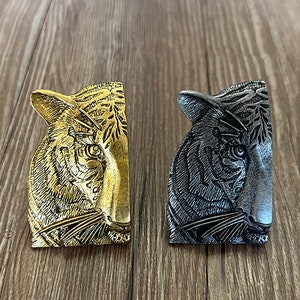 Puede incluir: Dos esculturas decorativas de cabeza de tigre. Una es dorada, la otra plateada. Ambas tienen tallas detalladas de la cara y el pelaje del tigre. Las esculturas se exhiben sobre una superficie de madera.