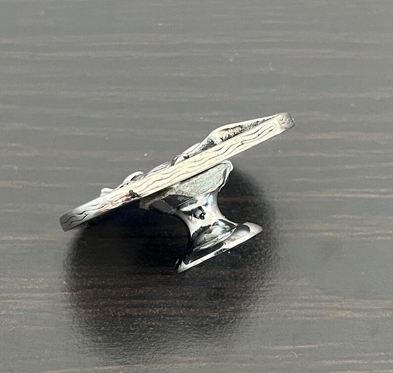 May include: A silver-colored, decorative cabinet knob with a detailed, textured design. The knob features a stylized, curved top and a pedestal base. The metal has a polished, reflective surface, and the design appears to be of a natural, organic form.