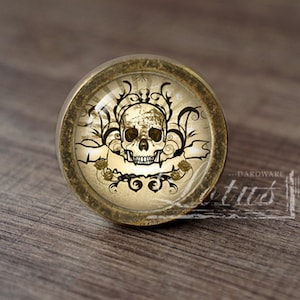 Puede incluir: Un pomo de armario redondo con diseño de calavera. El pomo presenta un cristal abovedado transparente sobre un fondo color crema con una calavera, detalles florales y una pancarta. El pomo tiene un borde metálico dorado.
