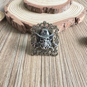 Può includere: Fibbia per cintura in metallo argentato con un teschio che indossa un cappello da cowboy e tiene due pistole. Il teschio è all'interno di una cornice rettangolare decorativa con dettagli floreali. Un accessorio unico.