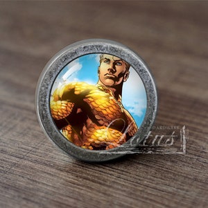 Könnte beinhalten: Ein silberner Knopf mit einer runden Glasplatte, die ein Comicbild von Aquaman zeigt, einem Superhelden mit orangefarbenen und goldenen Schuppen vor blauem Himmel.