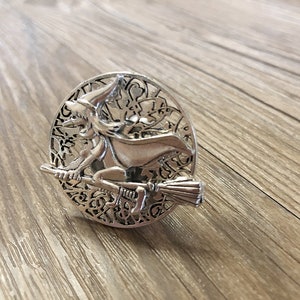 Op de afbeelding: Zilverkleurig metalen ornament met een heks die op een bezem vliegt, omringd door een cirkelvormig ontwerp met ingewikkelde details.