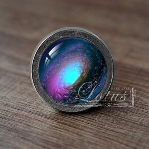 Puede incluir: Un pomo redondo de color plateado con un diseño de galaxia. La galaxia se representa en tonos de azul, morado y rosa.