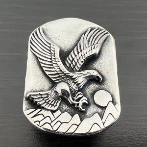 May include: A silver-toned rectangular ring featuring a detailed relief of an eagle in flight. The design includes mountains, a full moon, and intricate feather detailing. The ring has a slightly distressed finish.
