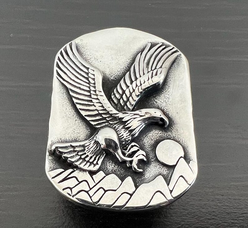 May include: A silver-toned rectangular ring featuring a detailed relief of an eagle in flight. The design includes mountains, a full moon, and intricate feather detailing. The ring has a slightly distressed finish.