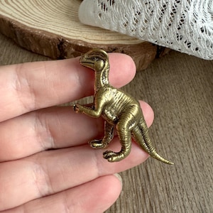 Könnte beinhalten: Kleine, antik-messingfarbene Dinosaurierfigur. Der Dinosaurier steht mit ausgestreckten Armen. Die Figur hat eine detaillierte Textur und ist wahrscheinlich aus Metall. Der Hintergrund umfasst eine Holzoberfläche und weiße Spitze.