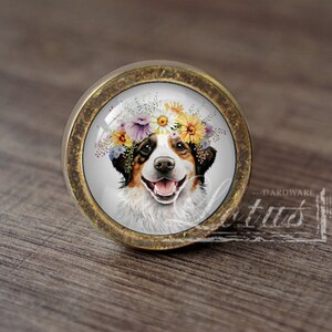 May include: A round, brass-coloured cabinet knob with a glass insert featuring a dog wearing a flower crown. The dog is white with brown and black markings and has a happy expression.