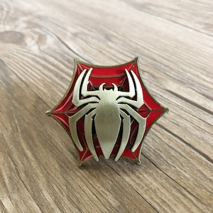 May include: A gold-coloured metal pin with a red and white spider web design. The spider is in the centre of the web and is also gold-coloured.