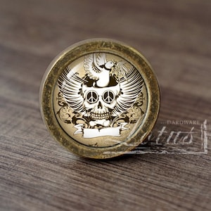 Könnte beinhalten: Runder Schrankknopf mit Totenkopf- und Friedenszeichen-Design. Der Knopf zeigt einen Totenkopf mit Friedenszeichen als Augen, Flügeln und einer Taube. Das Design ist in Weiß und Beige gehalten und befindet sich auf einer bronzefarbenen Metallbasis.