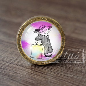 Pode incluir: Um botão de armário redondo, dourado, com um fundo colorido em estilo aquarela. No centro do botão, uma ilustração em preto e branco de uma pessoa usando um chapéu rosa e carregando uma sacola de compras.