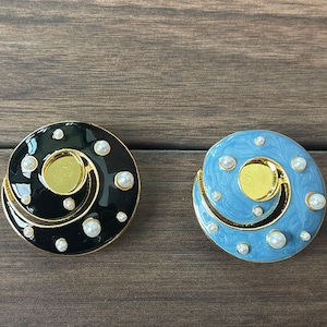 Könnte beinhalten: Zwei runde Broschen mit einem Spiraldesign. Eine Brosche ist schwarz mit weißen Perlen, die andere ist blau mit weißen Perlen.