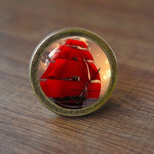 May include: A round, antique gold-toned cabinet knob with a glass insert featuring a red sailboat with white sails against a sunset sky.