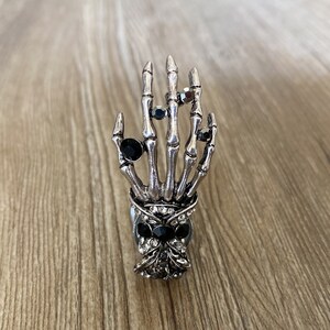 K&ouml;nnte beinhalten: Ein silberner Skeletthandring mit schwarzen Strasssteinen. Der Ring ist so gestaltet, dass er wie eine Skeletthand mit f&uuml;nf Fingern und einem Sch&auml;del an der Basis aussieht.