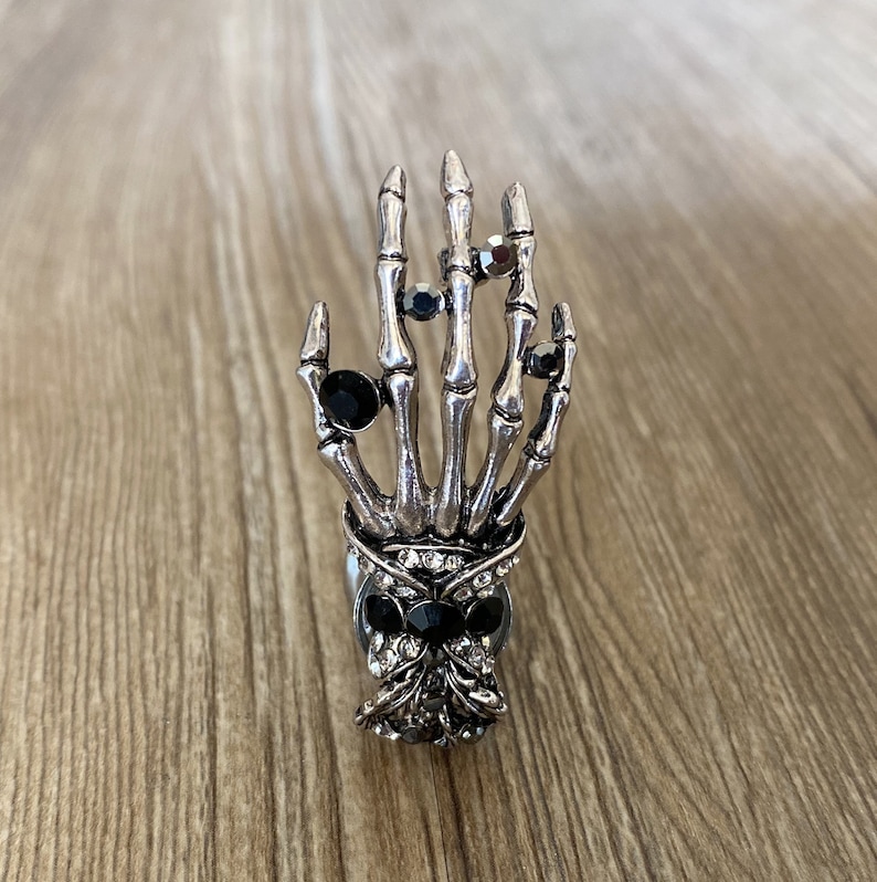 K&ouml;nnte beinhalten: Ein silberner Skeletthandring mit schwarzen Strasssteinen. Der Ring ist so gestaltet, dass er wie eine Skeletthand mit f&uuml;nf Fingern und einem Sch&auml;del an der Basis aussieht.