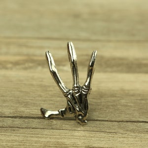 Puede incluir: Una mano de esqueleto plateada con tres dedos extendidos hacia arriba. La mano está hecha de metal y tiene un diseño detallado.