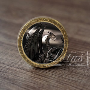 Op de afbeelding: Een ronde, gouden kastknop met een zwart drakenontwerp. De draak is gesilhouetteerd tegen een donkere achtergrond met een maan in de rechterbovenhoek.
