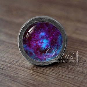 Może przedstawiać: Gałka meblowa w stylu galaktyki z srebrną metalową ramką i szklanym wstawką przedstawiającą wirującą mgławicę w odcieniach fioletu, błękitu i bieli.